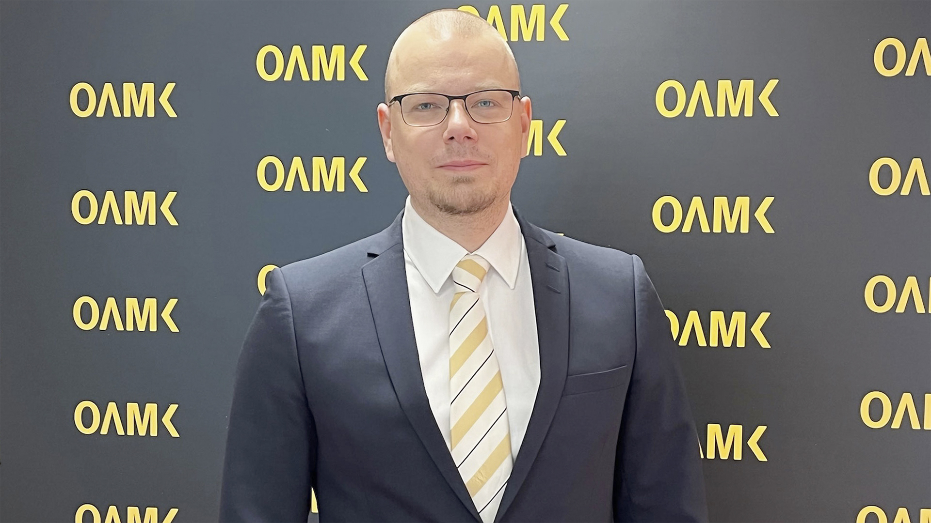 Pukuun pukeutunut mies seisoo Oamk-logolla varustetun kuvausseinän edessä.