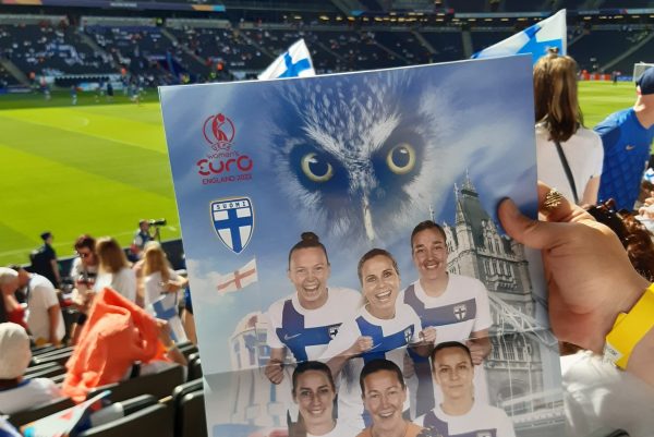 Valokuva jalkapallokatsomosta, jossa henkilö pitää kädessään Suomen naisten jalkapallojoukkueen kuvaa.