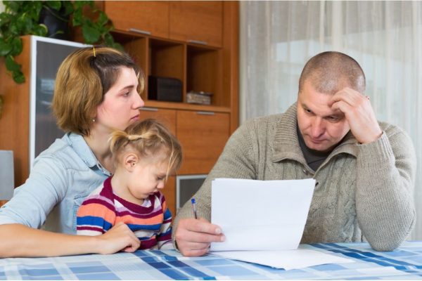 Valokuvassa yksilapsinen perhe istuu pöydän ääressä huolestuneen näköisenä ja katsoo paperia kädessään.